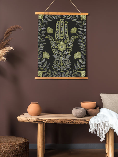 Botanical Hamsa wall tapestry featuring intricate sage green and lavender design on black background, showing a protective hand symbol surrounded by leaf motifs and floral elements, hanging from wooden dowels with natural twine.